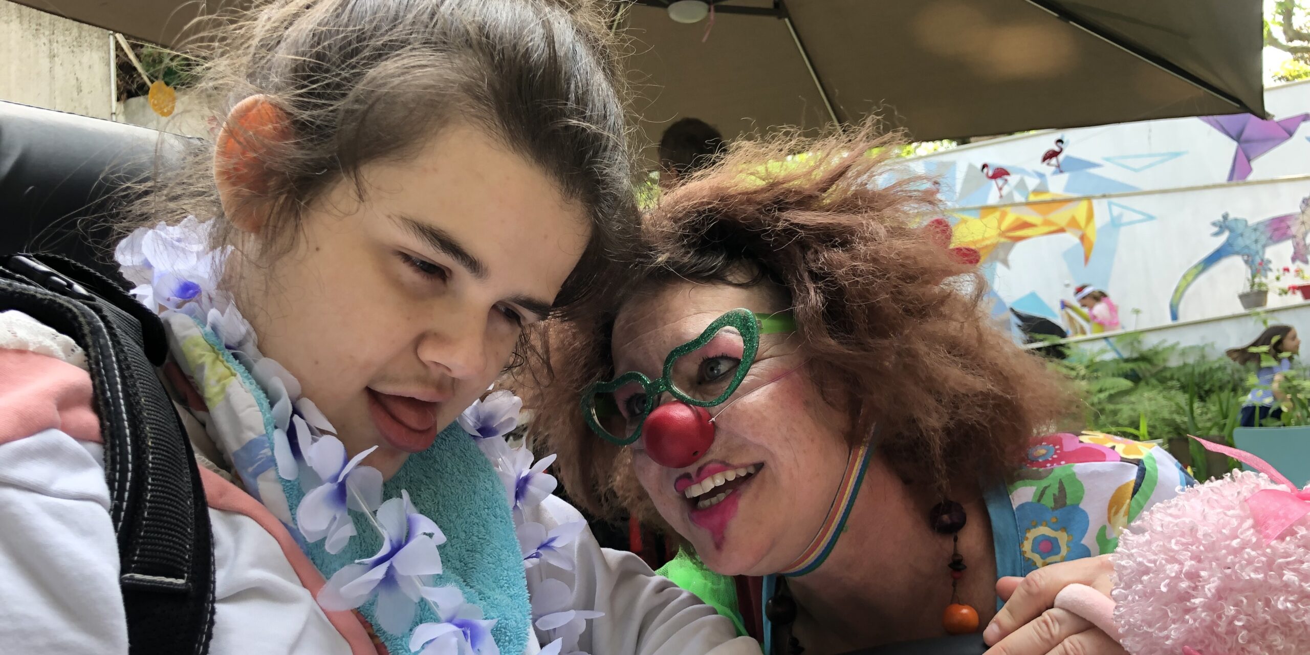 Une femme clown chuchote à l'oreille d'une jeune femme polyhandicapée, très attentive
